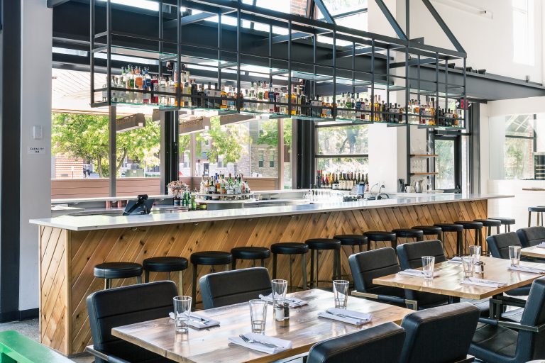 Restaurant bar with overhead bottle storage, wood panel front and seating with black chairs and wood tables in front of the bar