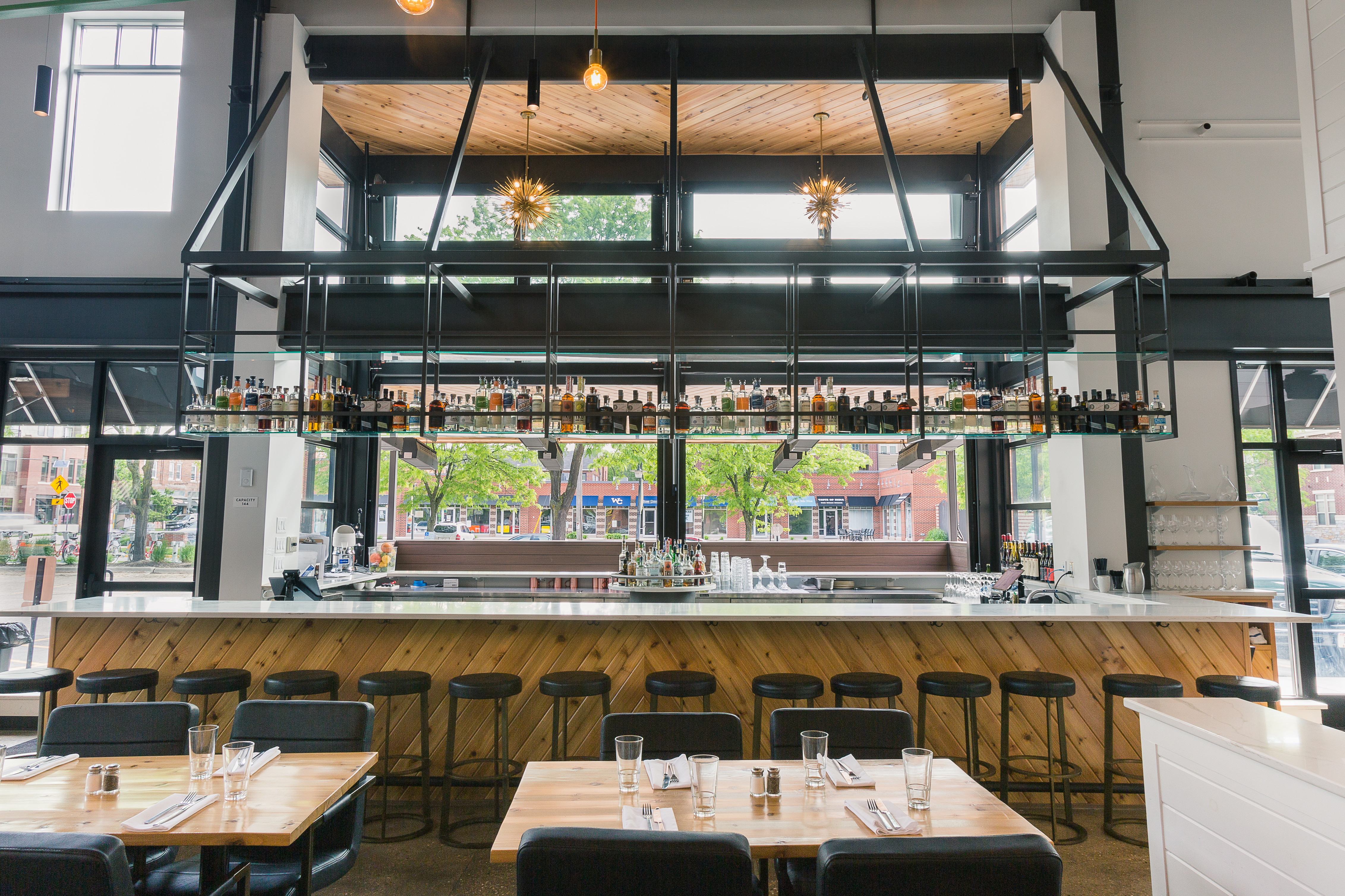 Wide shot of restaurant double sided bar that faces the inside seating area and outdoor patio, featuring metal overhead bottle storage, a wood facing bar with granite countertops