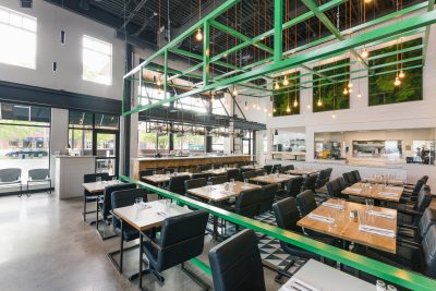 Wide shot of restaurant seating area with wood tables, black chairs, and green metal grid with hanging lights from the exposed ceilings