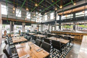 Restaurant seating area with green metal gird hanging from exposed ceiling with black chairs, concrete floors and wood table top tables with view of the bar