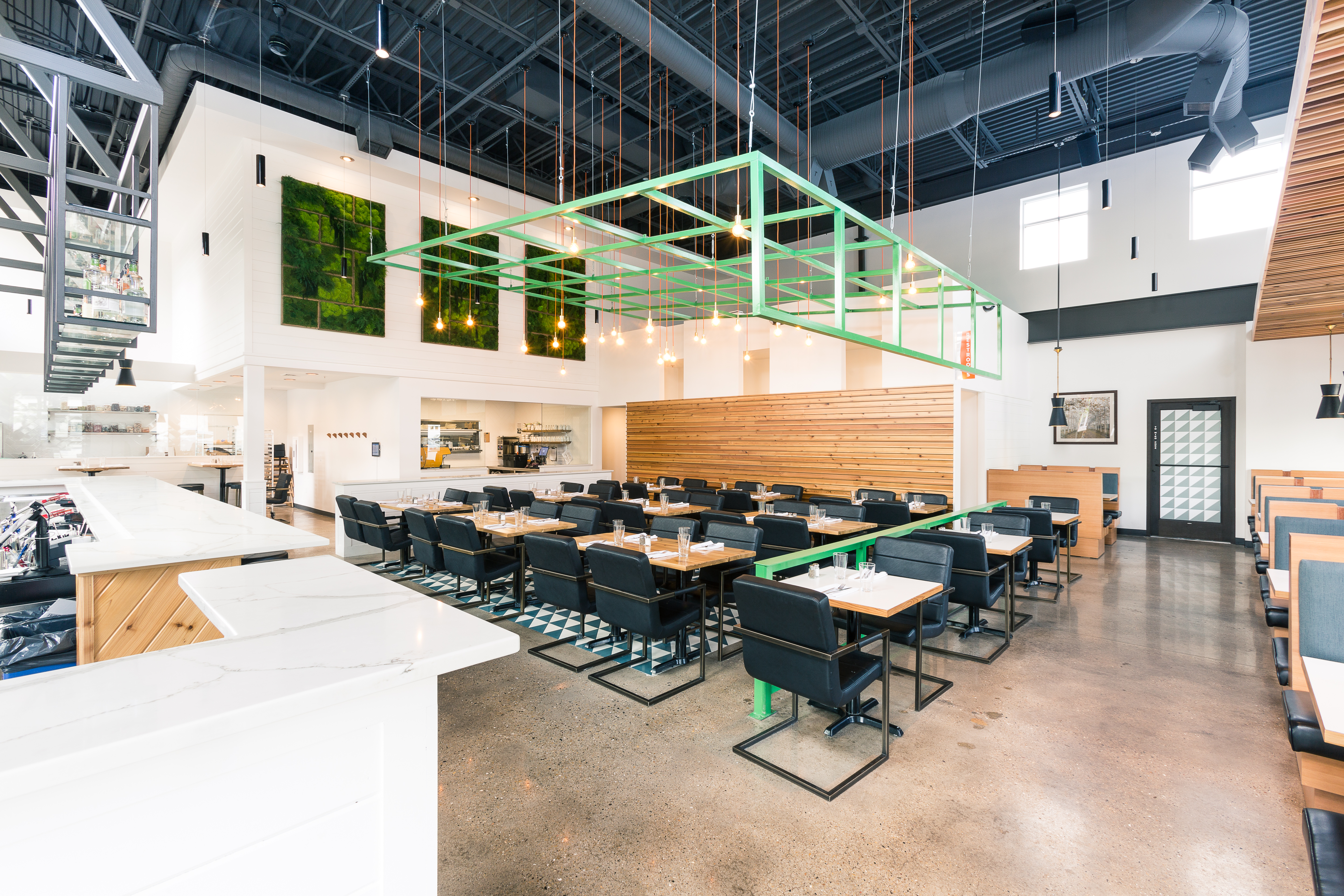 Restaurant seating area with green metal gird hanging from exposed ceiling with black chairs, concrete floors and wood table top tables