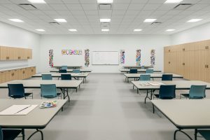 A brightly lit art classroom with several tables and chairs arranged neatly. Art supplies, including paint palettes and paper, are on the tables. Colorful posters and student artwork are displayed on the walls. Cabinets line the walls for storage.