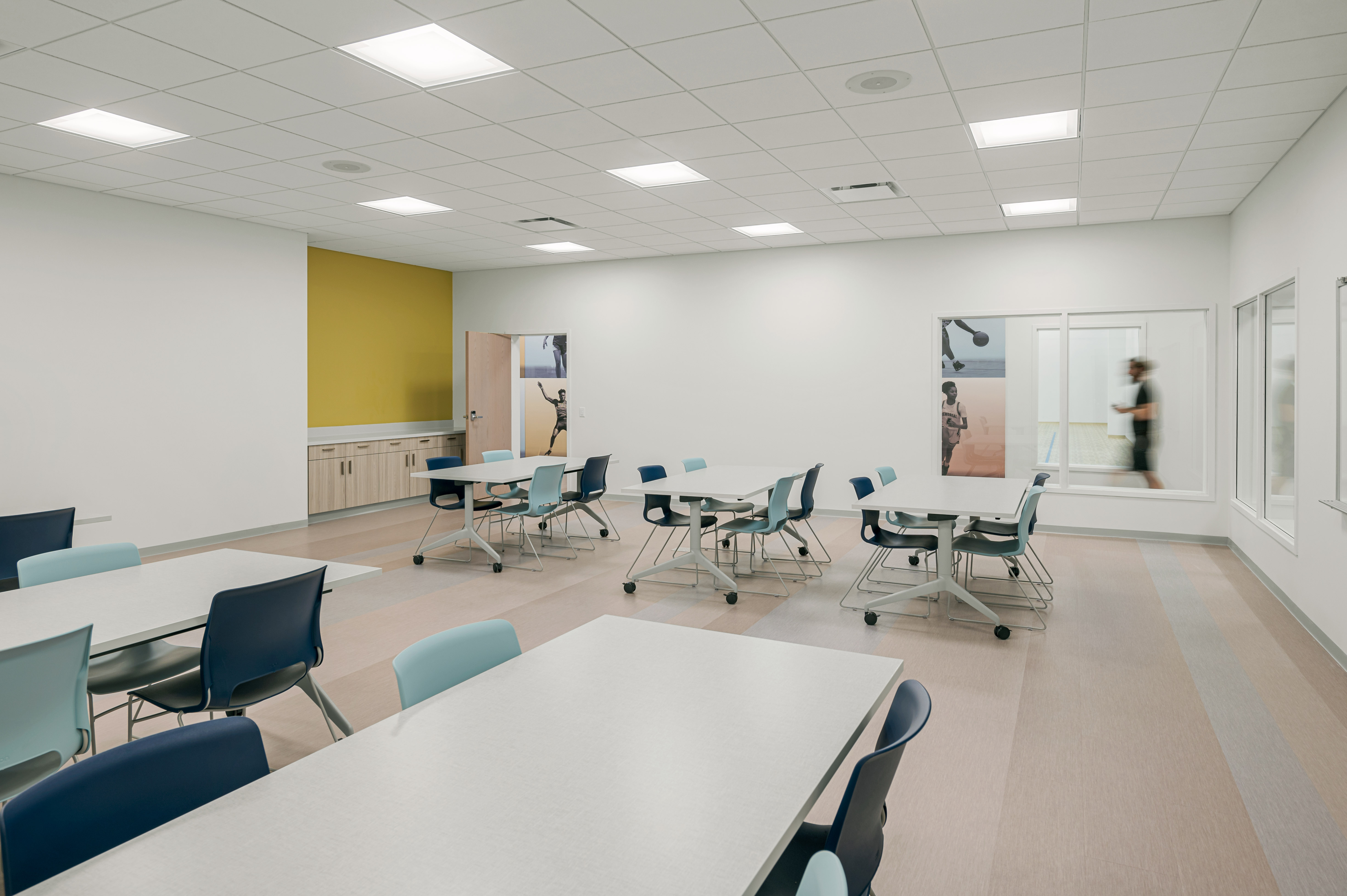 A clean, modern classroom with white walls and a yellow accent wall at the back. Several tables and chairs are arranged in rows. A door and windows on the right side reveal a hallway where a blurry figure is walking. The ceiling has recessed lighting.