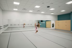 A young person in a pink outfit is dancing energetically in a well-lit dance studio with large mirrors along one wall. A person, her mother stands near the hallway window observing. The studio features ballet barres and wooden cabinets.