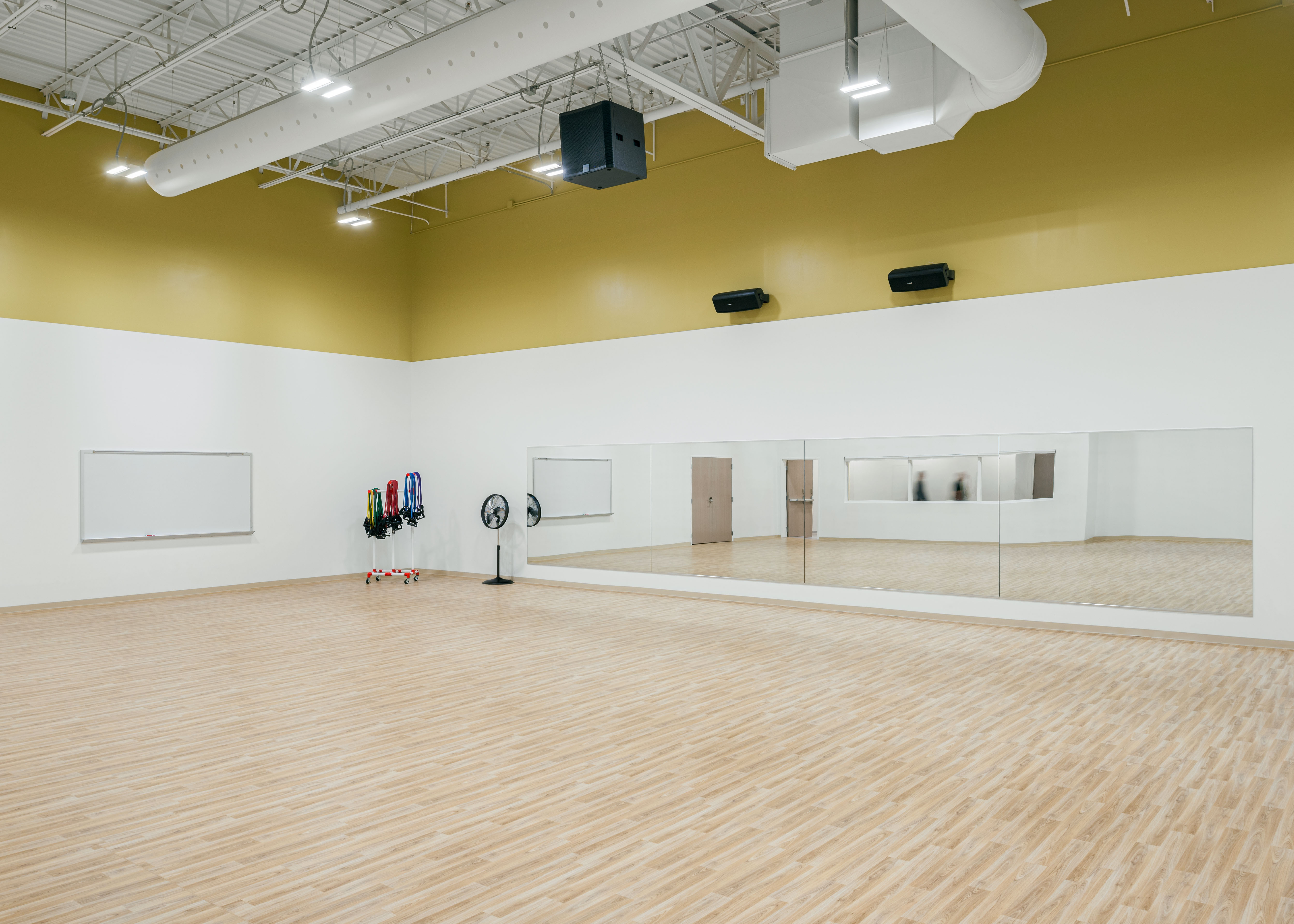 A spacious, empty dance studio with wooden flooring, white walls, and a high ceiling. Large mirrors are mounted on one wall, with a fan and various resistance bands hanging on the wall in the corner. The room is well-lit from overhead lights.