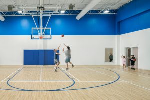 Two people playing basketball in a modern indoor court with blue and white walls. One player jumps to shoot the ball, while the other attempts to block. In the background, a couple with a child and another person holding a basketball observe from the entrance.