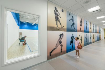 A young girl holding a basketball stands in a hallway, gazing at a wall adorned with large photos of athletes. Through a window to her left, a man is seated on a chair inside a basketball court, holding a basketball.