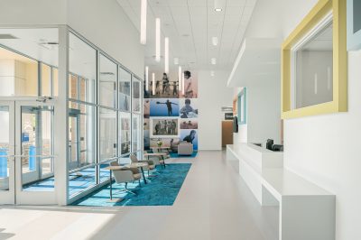 A modern, bright lobby area featuring a glass entrance, minimalistic white interior, and a vivid blue carpet. The space is furnished with contemporary chairs and decorated with a large photo collage on the wall. Fluorescent lights hang from the high ceiling.