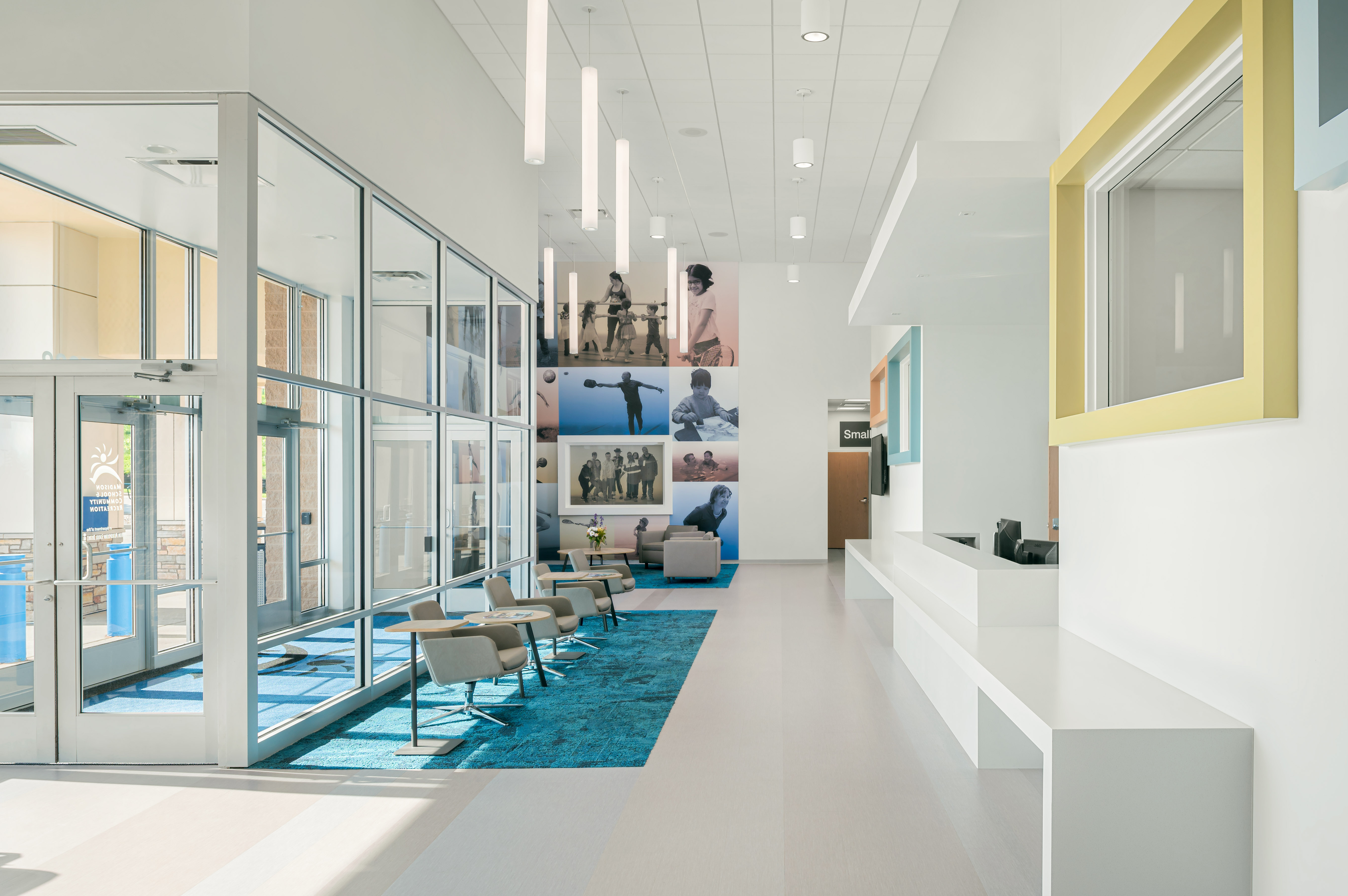 A modern, bright lobby area featuring a glass entrance, minimalistic white interior, and a vivid blue carpet. The space is furnished with contemporary chairs and decorated with a large photo collage on the wall. Fluorescent lights hang from the high ceiling.