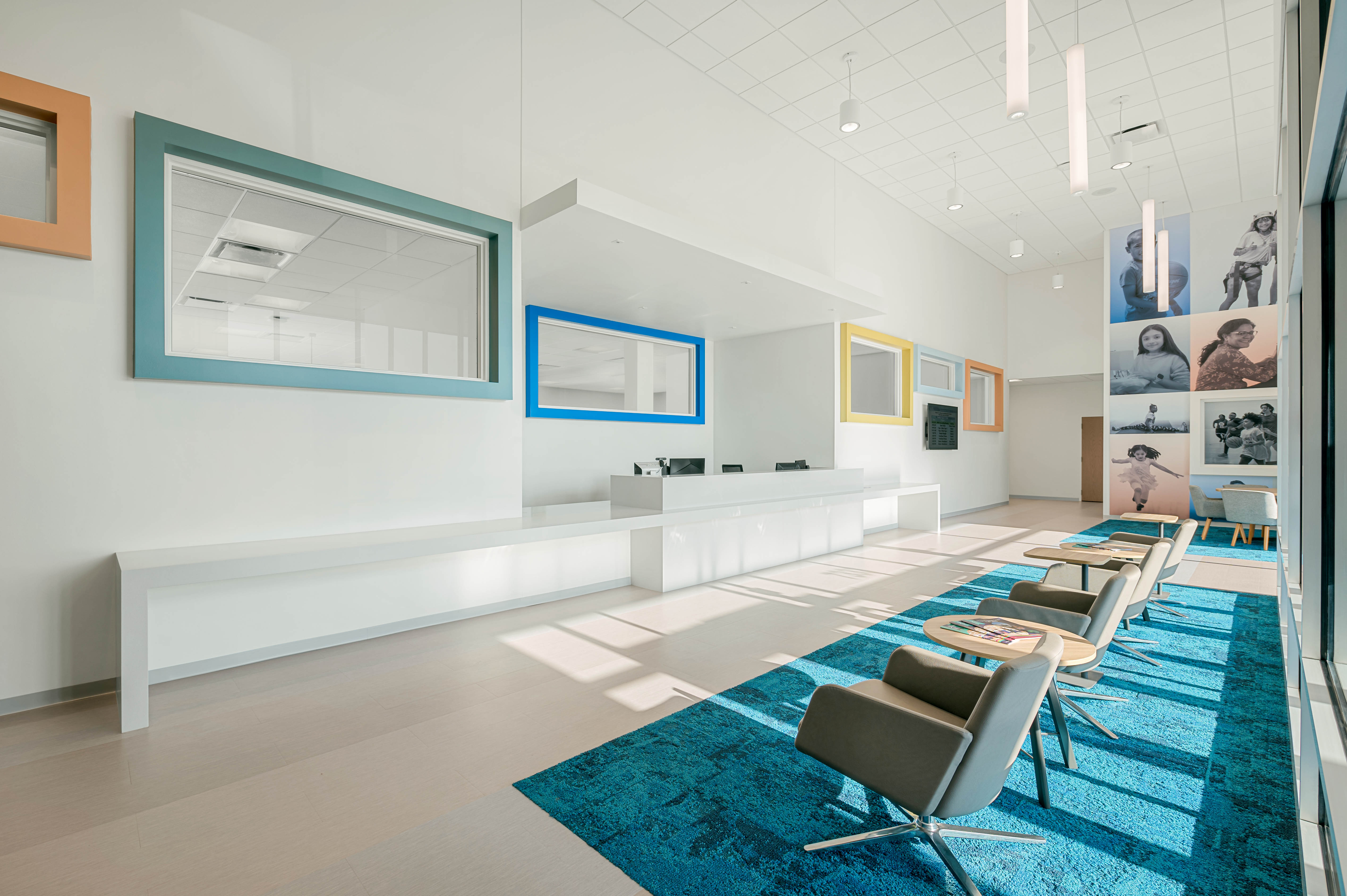 A modern, brightly lit waiting area with a white counter and reception desk. The room features high ceilings, large windows, and blue and yellow framed wall accents. Comfortable gray chairs and a blue patterned rug add warmth. Photos on the wall display people.