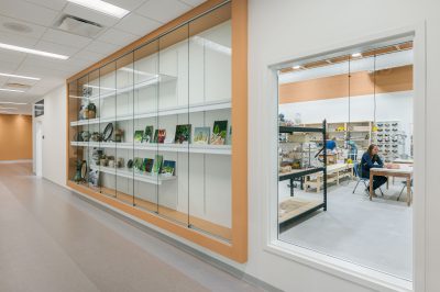 A hallway with display shelves on the left showcasing various artworks. On the right, a large window reveals a pottery studio with shelves of ceramics and a person working at a table. The walls are painted in a warm beige tone.