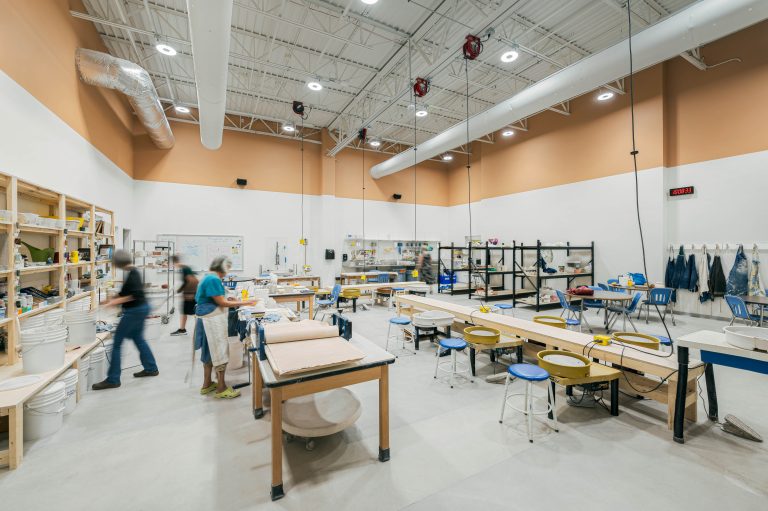 A spacious, well-lit ceramics workshop filled with various equipment and supplies. Several people are engaged in different activities around worktables. Shelves with tools and materials line one wall, and pottery wheels are set up near the center of the room.