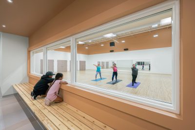 Three people are practicing yoga inside a spacious studio, viewed from the perspective of a the hallway with a large window. Two children and an adult are watching them from outside while sitting on a wooden bench. The yoga practitioners are balancing on mats.