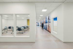 A modern hallway with a paneled ceiling and light flooring. On the left, a classroom with desks and chairs is visible through a large window. On the right, a person is walking past a wall adorned with large artistic images.