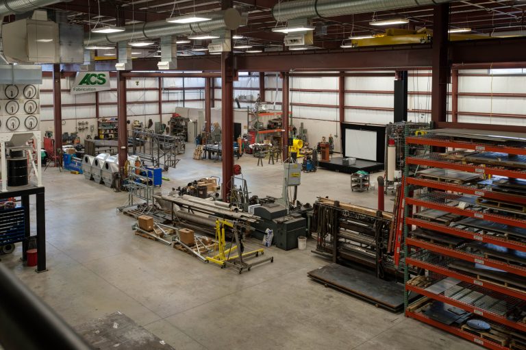 spacious industrial warehouse with metal shelves, machinery, and fabrication tools. Workers in safety gear operate equipment. Large overhead ducts are visible, and the space is well-organized for manufacturing tasks.