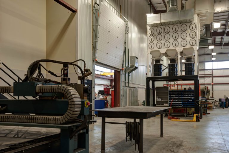 Industrial workshop with machinery, including a large metal table and equipment, in a spacious, well-lit area. A ceiling-high dust collection system with multiple filters is in the background, adjacent to open garage-style doors.