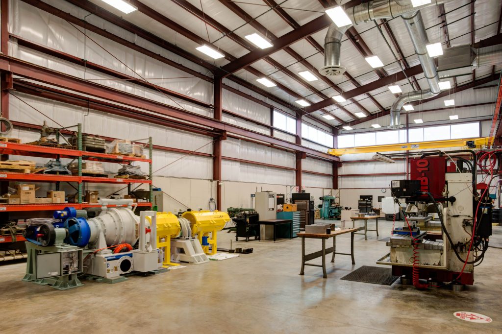 A spacious industrial workshop with high ceilings, featuring various machinery and equipment. Metal shelves with tools and materials line the walls. The floor is clean and the area is well-lit with overhead lighting.