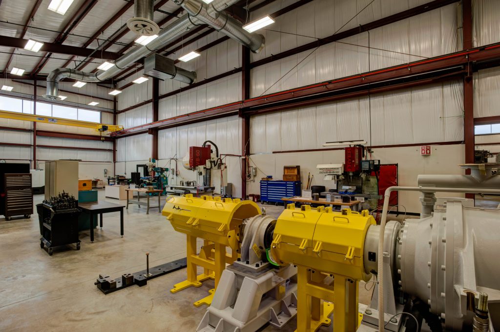 Interior of an industrial workshop with a high ceiling, showcasing various equipment. Prominent yellow machinery in the foreground, workbenches, tools, and metal pipes.