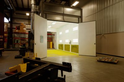 A large industrial paint booth with open doors revealing a brightly lit interior and yellow-colored floor. The surrounding area has shelves with various equipment and tools. The workshop has high ceilings and steel walls.