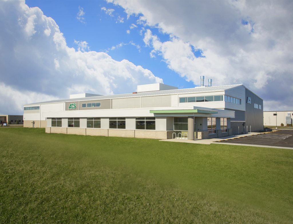 industrial building with a silver and white facade is set against a partly cloudy sky. The building features large windows and a green logo on the side.