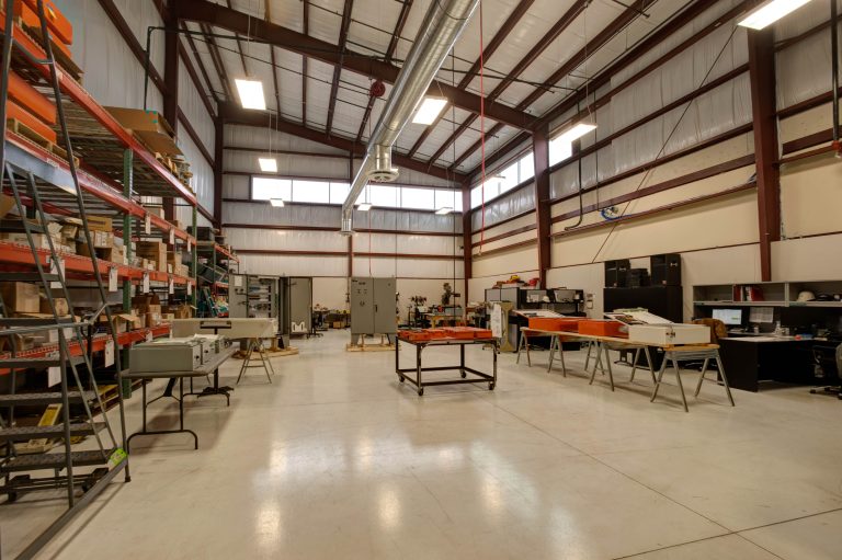 spacious industrial workshop with high ceilings and large windows. Metal shelves line the walls filled with supplies. Several workbenches and equipment are scattered throughout the polished concrete floor, illuminated by overhead lighting.