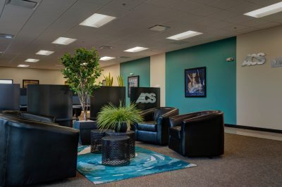 office lobby featuring black leather chairs arranged around small round tables on a patterned rug. Green plants add a touch of nature, and framed art decorates the walls.