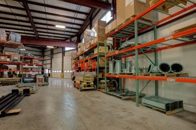 warehouse with high metal shelves filled with boxes and industrial equipment. Pallets of materials are stacked neatly, and a loading dock with large closed doors is visible in the background.