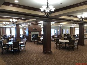 A spacious dining area with numerous tables and chairs under elegant chandeliers. The room has carpeted flooring and wooden columns with ceiling beams. Large windows are seen in the background, and a fireplace in the center.