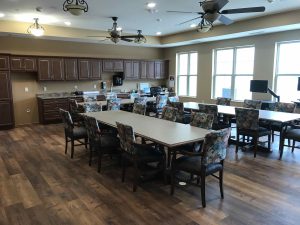 A well-lit room with wooden flooring featuring three long tables surrounded by chairs with floral upholstery. The room has several ceiling fans, large windows, and a kitchen area with dark wooden cabinets, a sink, and various kitchen appliances.