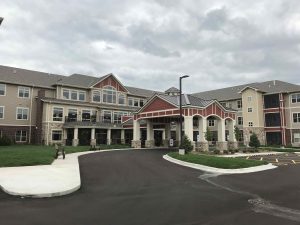 A large, modern senior living complex with three floors and a mix of brick and siding exterior. The entrance is accentuated by columns and a covered drop-off area. The grounds feature landscaped grass, a paved driveway, and a parking area. Cloudy skies overhead.