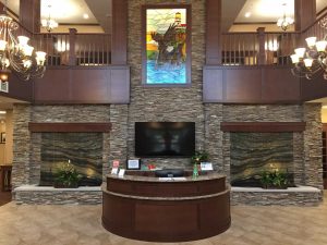 The image shows a luxurious senior living facility lobby featuring a round reception desk made of granite and wood. Behind the desk is a wall with stacked stone tiles, a flat-screen TV, and a tall stained glass window with a lighthouse design. Chandeliers hang from the ceiling.