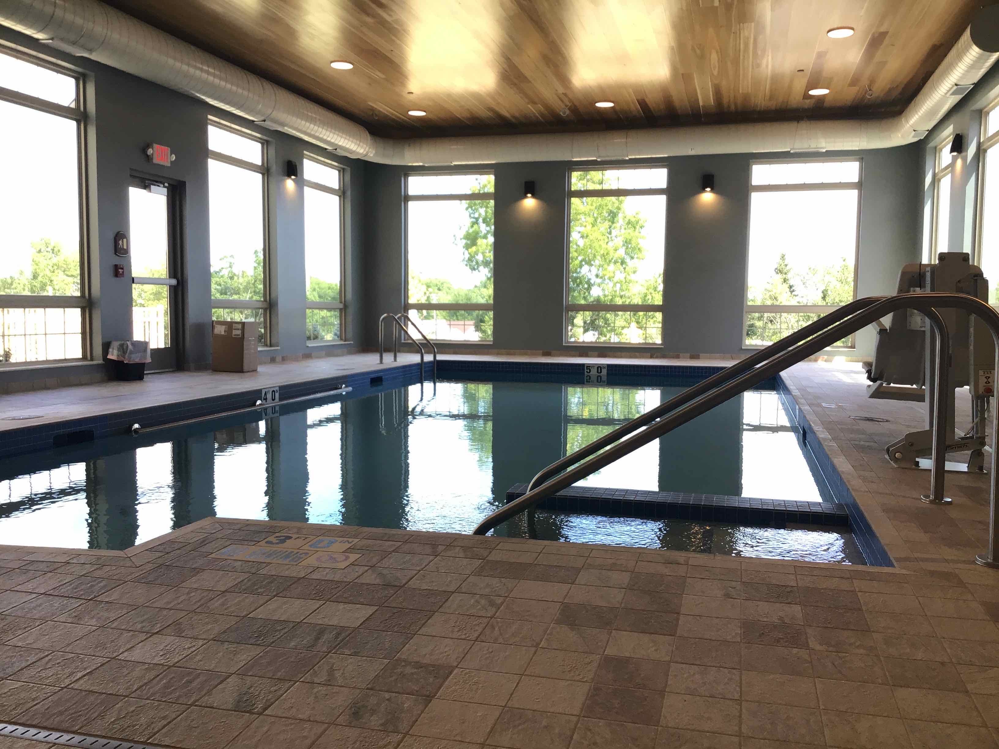 Indoor swimming pool in a well-lit room with large windows. The pool has metal handrails and a tiled deck. In the background, trees are visible through the windows. The ceiling is wooden, and there's a lifeguard chair and emergency equipment near an exit door.