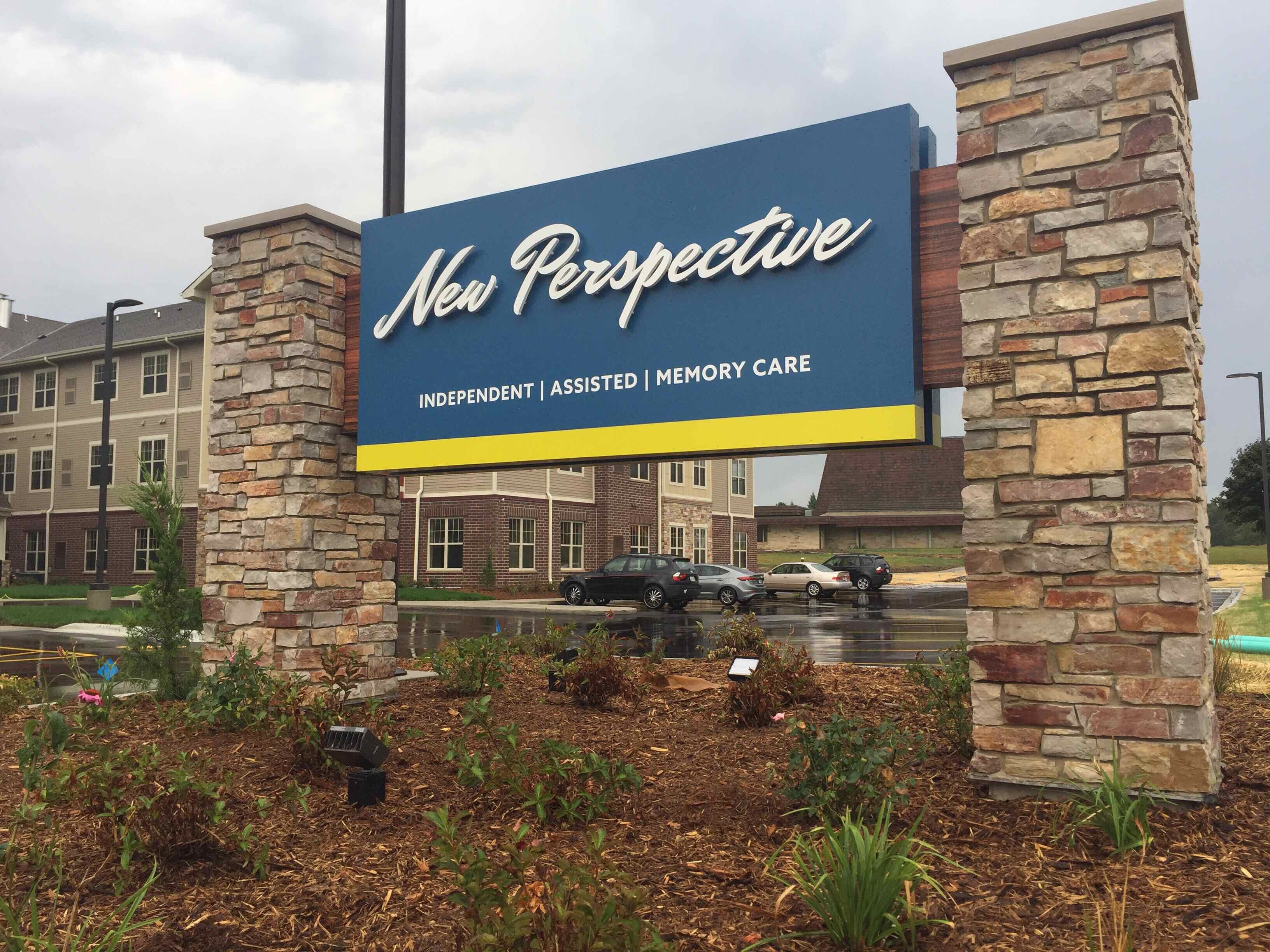 A sign for "New Perspective" assisted living facility stands between stone pillars. The sign has text reading "INDEPENDENT | ASSISTED | MEMORY CARE." In the background, there is a brick building and a parking lot with cars.