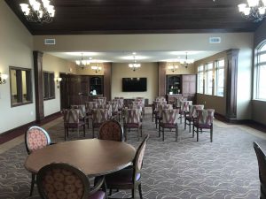 A theater room with a large TV mounted on the front wall, rows of patterned chairs facing the TV, round tables with chairs near the back, chandeliers hanging from the ceiling, and large windows on the right side.