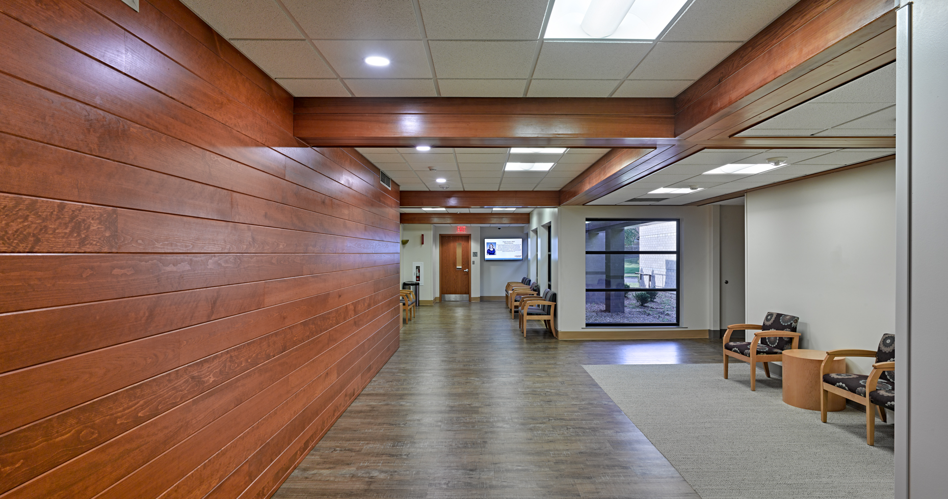 A hallway in a building with wooden wall panels and a wooden floor. The ceiling has recessed lighting. There are several chairs with small tables arranged against the walls, and a window on the right side showing a view outside.