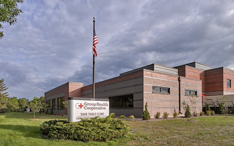 A red and grey brick building with a large sign in front reading "Group Health Cooperative Sauk Trails Clinic Complementary Medicine Center". The building is surrounded by a grassy lawn and trees, with a partly cloudy sky in the background.