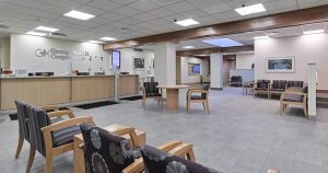 A spacious, well-lit waiting room with multiple seating areas, including cushioned chairs and small tables. The sign "Group Health Cooperative" is visible above the reception desk. The walls are decorated with various artworks, creating a welcoming atmosphere.