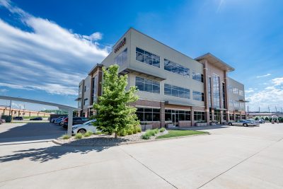 A modern, multi-story office building with large windows. The surrounding area includes a parking lot with several cars and a few trees