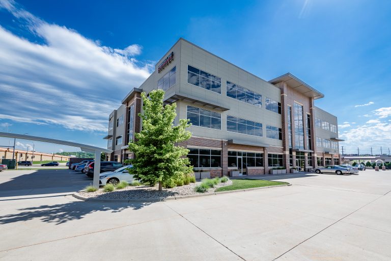 A modern, multi-story office building with large windows. The surrounding area includes a parking lot with several cars and a few trees