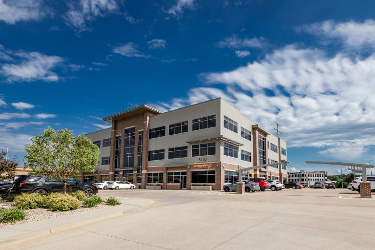 Office building with three floors under a clear blue sky, surrounded by parked cars. The building has large windows and a modern facade, with a few clouds visible in the sky.