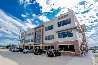 A modern, three-story office building with large windows, surrounded by a parking lot with several cars. The building has a mix of beige and brick exterior.