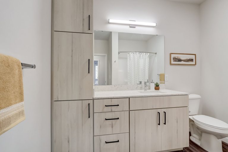 A modern bathroom with a light wood vanity, white countertop, large mirror, and a white toilet. A yellow towel hangs on the left wall, and a small framed artwork is above the toilet.