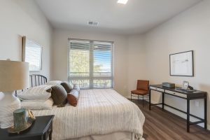 A cozy bedroom with a large window, a bed with striped bedding and multiple pillows, and a dark wood nightstand with a lamp. A minimalist desk with a chair is placed against the opposite wall, featuring framed artwork above it.