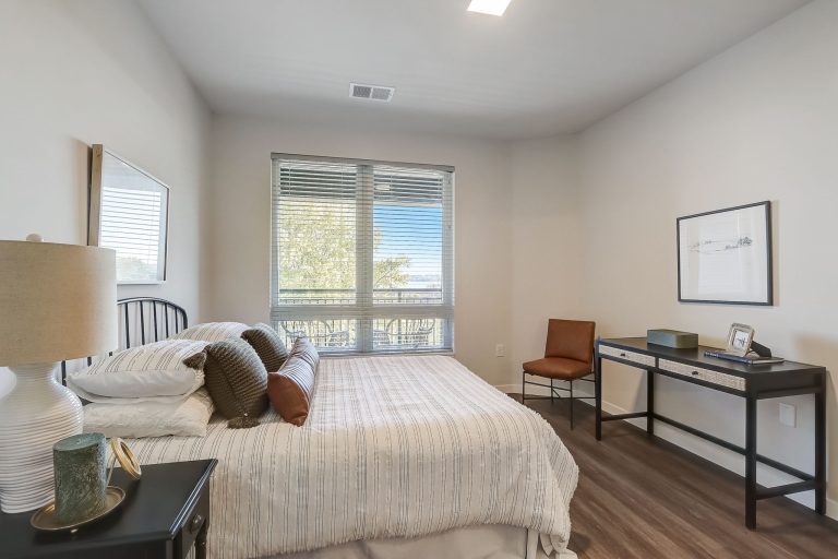 A cozy bedroom with a large window, a bed with striped bedding and multiple pillows, and a dark wood nightstand with a lamp. A minimalist desk with a chair is placed against the opposite wall, featuring framed artwork above it.