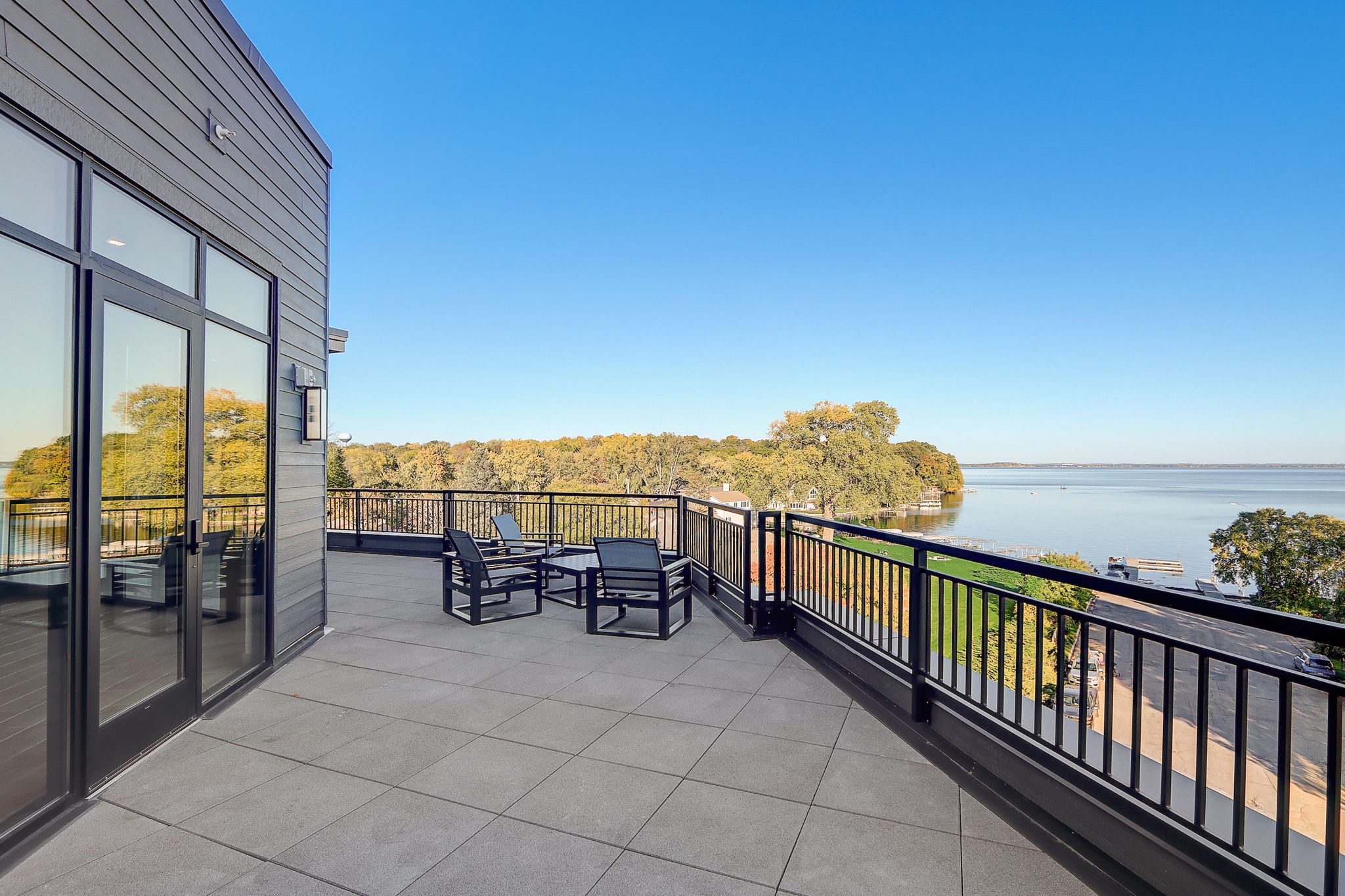 A spacious rooftop balcony with outdoor seating, overlooking a scenic view of a lake surrounded by trees and greenery. The balcony features glass doors leading inside and is bordered by a black metal railing.