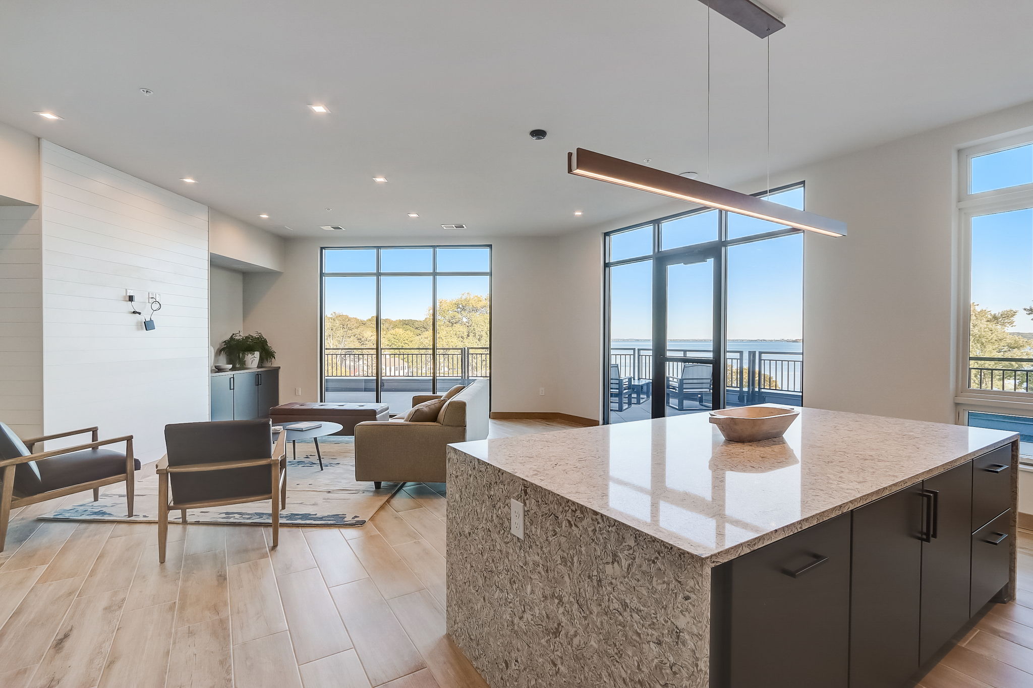 A modern, open-concept apartment community room with large windows offering a scenic view of a lake, natural wood flooring, and a kitchen island with a stone countertop