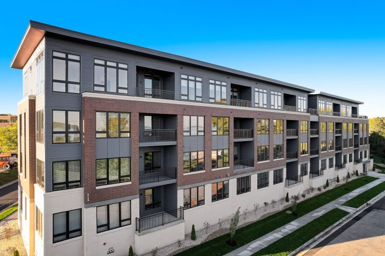 Exterior of a modern four-story apartment building with a mix of brick and siding exterior, large windows, and private balconies, set against a clear blue sky