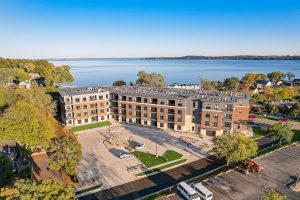 Exterior aerial view of a modern apartment complex with brick and siding exterior, situated near a large lake, surrounded by trees and residential houses