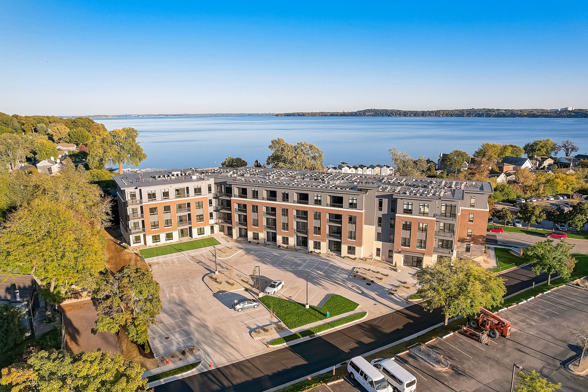 Exterior aerial view of a modern apartment complex with brick and siding exterior, situated near a large lake, surrounded by trees and residential houses