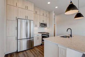 Modern kitchen with light wood cabinets, stainless steel appliances including a French door refrigerator, and a large island featuring a sink and black faucet. Pendant lights hang above the island